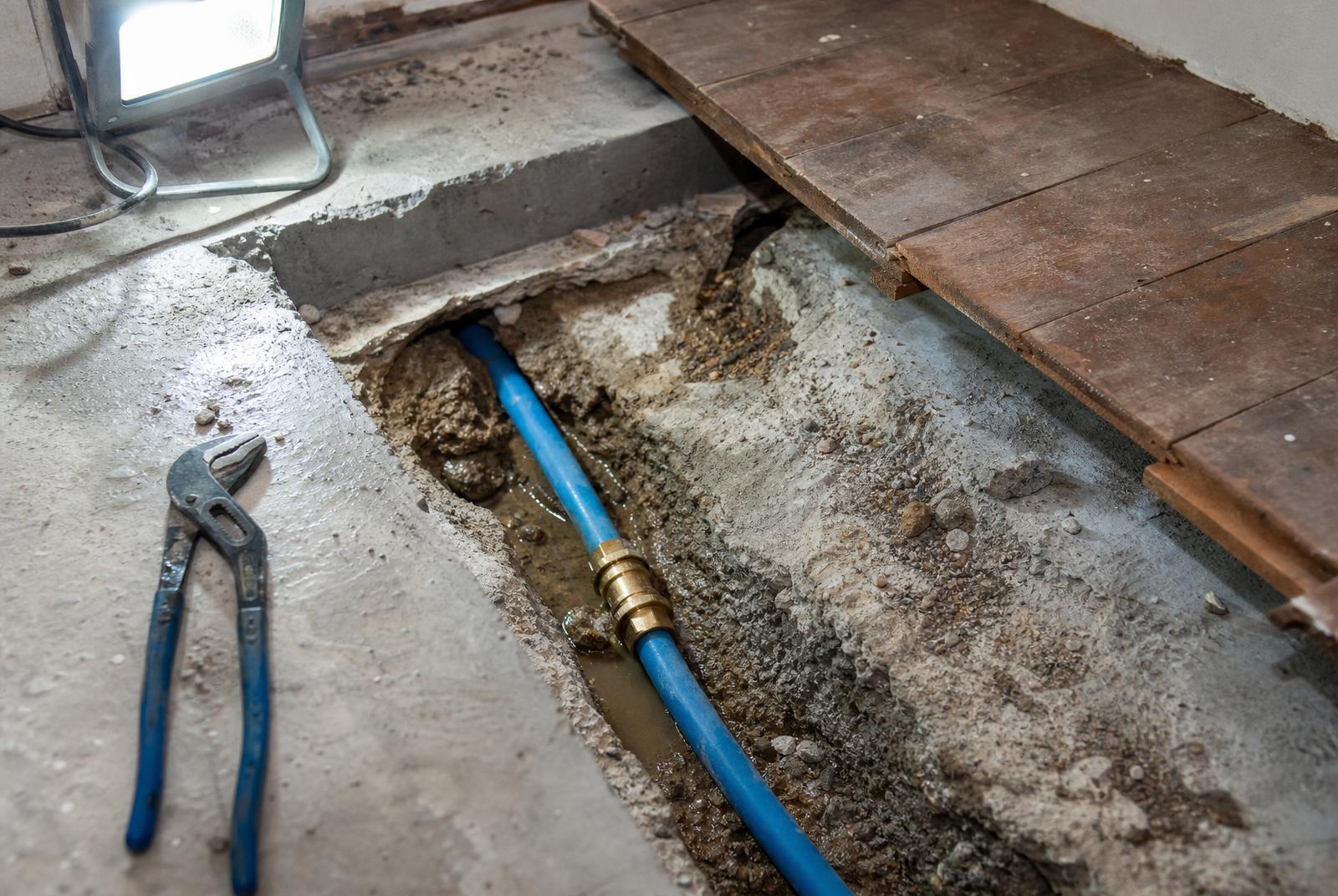 Repaired blue mains water pipe with a new brass coupling in an opened floor under work-lamp light